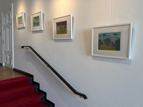 Vier bunt gerahmte Bilder hängen an einer weißen Wand neben einer Treppe mit rotem Teppich.