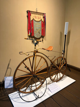 Historisches Fahrrad mit großen Rädern und ledernem Sattel, dahinter ein bannerähnliches Schild mit Verzierungen.