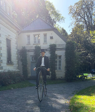 Mann in Frack fährt ein Hochrad auf einem gepflasterten Weg vor einem historischen Gebäude im Park.