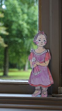 Papierfigur eines Mädchens in rosa Kleid mit Puppe, im Fenster vor einem grünen, unscharfen Hintergrund.