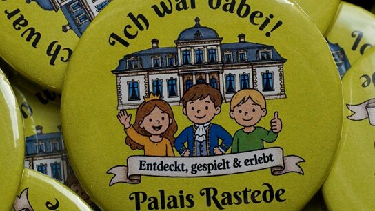 Bunte Ansteckknöpfe mit der Aufschrift „Ich war dabei! Palais Rastede“ und einer Illustration von drei Kindern.