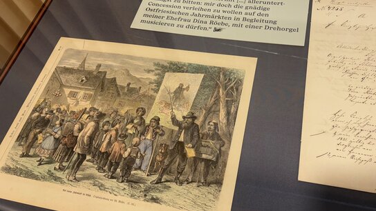 Eine farbenfrohe Lithografie zeigt eine Gruppe von Menschen, die sich um einen Redner versammeln, mit einer Landschaft im Hintergrund.