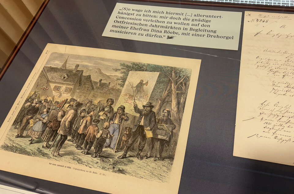 Eine farbenfrohe Lithografie zeigt eine Gruppe von Menschen, die sich um einen Redner versammeln, mit einer Landschaft im Hintergrund.