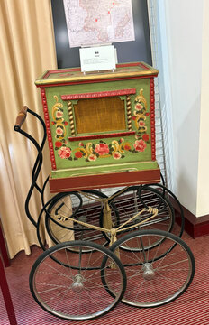 Bunt verzierter Straßenmusikautomat auf einem Wagen mit großen Rädern, im Hintergrund eine Landkarte.