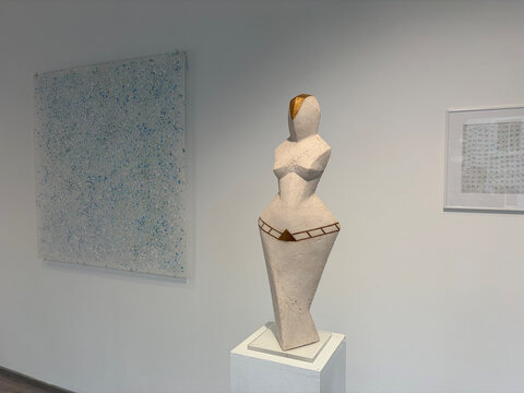 Skulptur einer weiblichen Figur in einem modernen Ausstellungsraum, umgeben von abstrakten Gemälden.