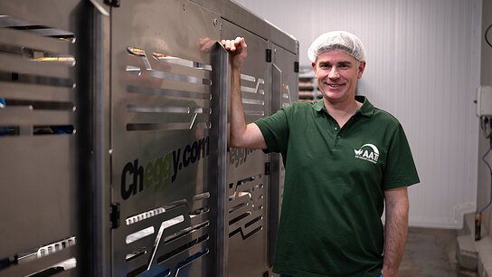 Mann in grüner Arbeitskleidung und Haarnetz steht lächelnd neben einer silbernen Maschinenreihe in einem modernen Arbeitsumfeld.