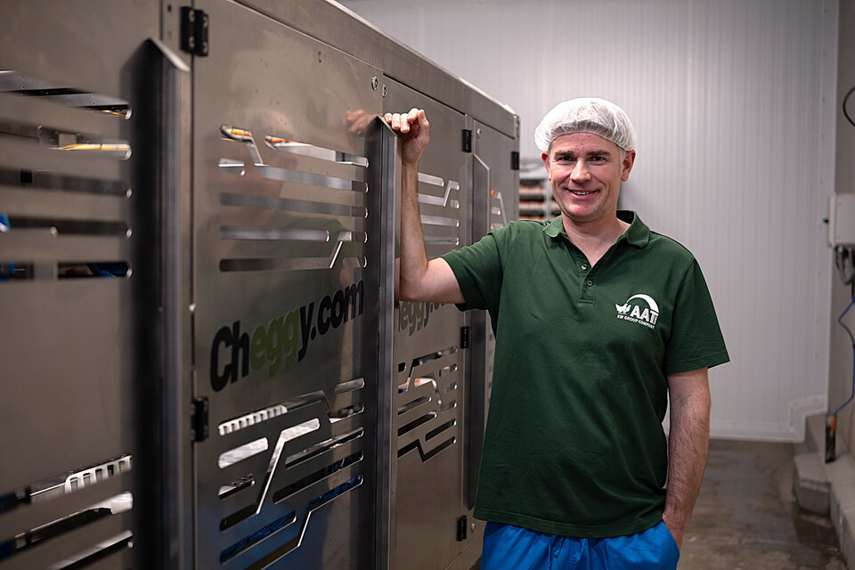 Mann in grüner Arbeitskleidung und Haarnetz steht lächelnd neben einer silbernen Maschinenreihe in einem modernen Arbeitsumfeld.