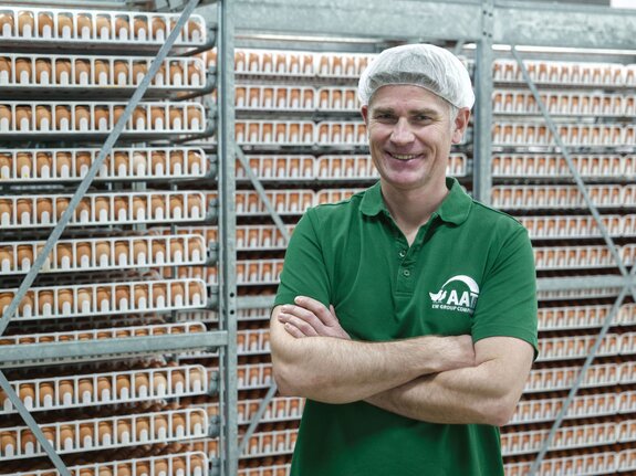 Lächelnder Mann in grünem T-Shirt steht vor Regalen mit Eiern in einer Produktionshalle.