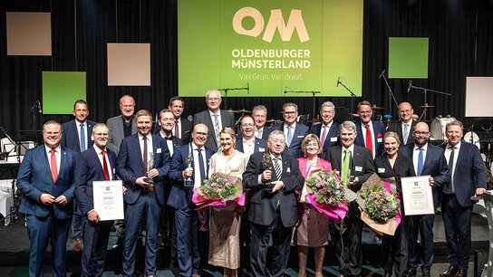 Gruppierung von 20 Menschen auf einer Bühne, die Urkunden halten und Blumen präsentieren, vor einem großen, beleuchteten Logo.