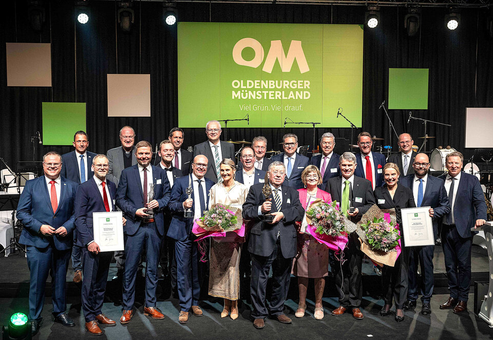 Gruppierung von 20 Menschen auf einer Bühne, die Urkunden halten und Blumen präsentieren, vor einem großen, beleuchteten Logo.