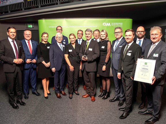 Gruppenfoto von zehn Personen bei einer Preisverleihung, einige halten Urkunden, vor einem grünen Hintergrund mit Logo.