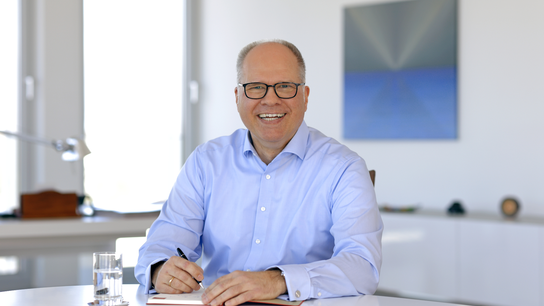 Michael Thanheisee in hellblauem Hemd sitzt an einem Tisch, hält einen Stift und notiert etwas. Heller, moderner Raum.