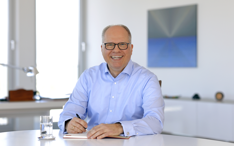 Michael Thanheisee in hellblauem Hemd sitzt an einem Tisch, hält einen Stift und notiert etwas. Heller, moderner Raum.