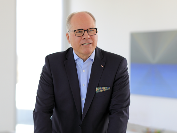 Michael Thanheiser steht lächelnd in einem modernen Raum mit großem Fenster und abstrakter Kunst im Hintergrund.