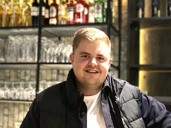 Mann mit lächelndem Gesicht steht an einer Bar, im Hintergrund Regal mit alkoholischen Getränken und Gläsern.