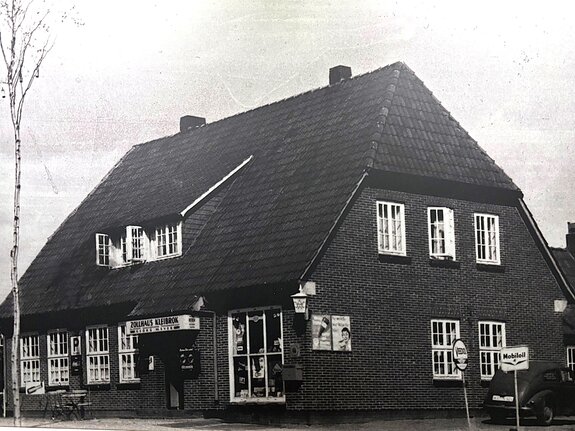 Zweistöckiges, dunkelrotes Fachwerkhaus mit Satteldach und vielen Fenstern, umgeben von Bäumen und einer Straße.