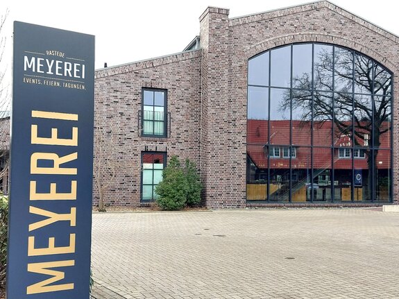 Moderne Eventlocation mit glasfrontiger Fassade, rustikaler Backsteinoptik und begrüntem Eingangsbereich.