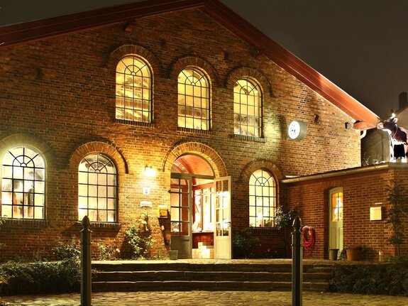 Historisches Backsteingebäude mit großen Fenstern, beleuchtet durch abendliches Licht, Eingangstür offen.