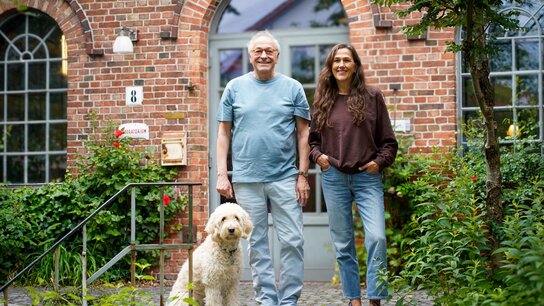 Pavel Möller-Lück und Barbara Schmitz-Lenders  stehen gemeinsam mit einem Hund vor einem Backsteingebäude, umgeben von Pflanzen und Blumen.