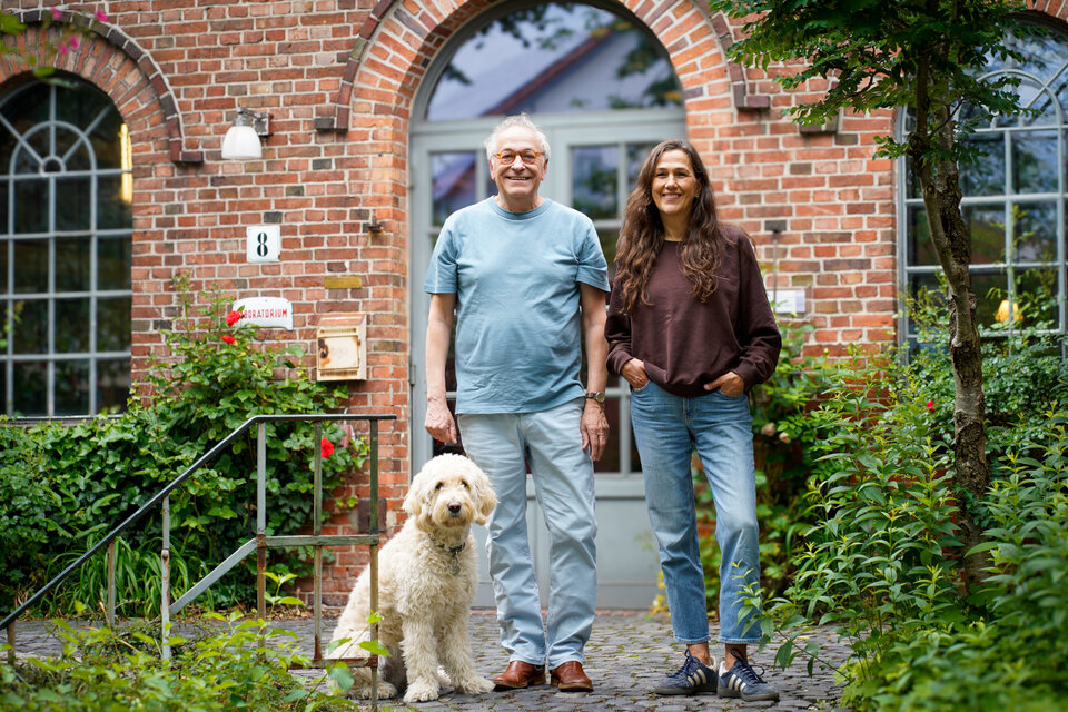 Pavel Möller-Lück und Barbara Schmitz-Lenders  stehen gemeinsam mit einem Hund vor einem Backsteingebäude, umgeben von Pflanzen und Blumen.