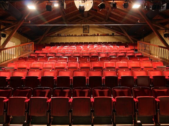 Leere Theaterbühne mit roten Sitzen und Holzkonstruktion, beleuchtet von oben.