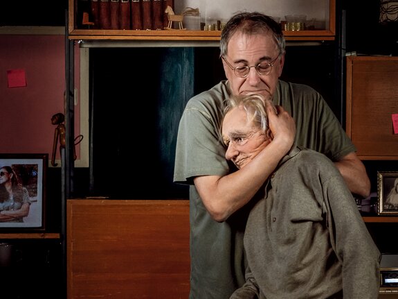 Ein Mann umarmt eine Puppe, die wie eine ältere Person aussieht, in einem Raum mit Bücherregalen und Familienfotos im Hintergrund.