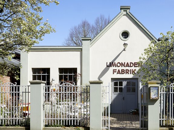 Ein modernes Gebäude mit dem Schriftzug "LIMONADE FABRIK", umgeben von Bäumen und einem Zaun.