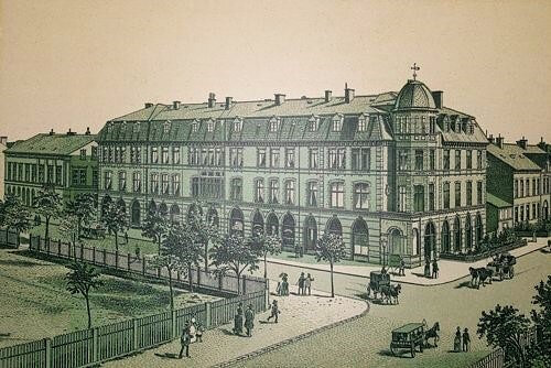 Historisches Gebäude mit prächtiger Fassade, umgeben von Bäumen und Straßen; Pferdewagen und Fußgänger zu sehen.