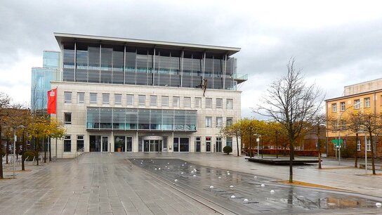 Moderner Gebäude mit Glasfassade, umgeben von Bäumen, einem Platz mit Wasserfontänen und grauen Steinplatten.