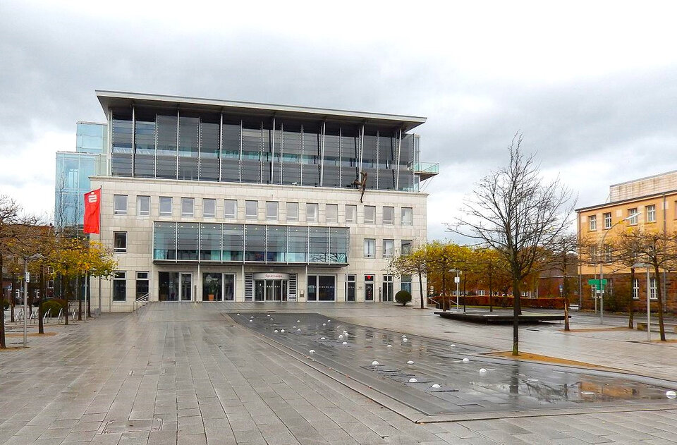 Moderner Gebäude mit Glasfassade, umgeben von Bäumen, einem Platz mit Wasserfontänen und grauen Steinplatten.