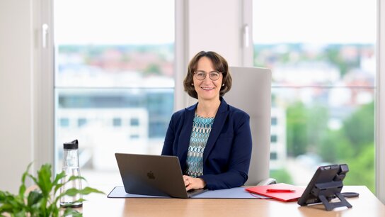 Frau mit Brille sitzt am Schreibtisch, arbeitet am Laptop, Blick aus dem Fenster auf eine Stadtlandschaft.