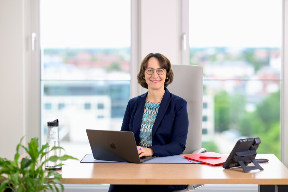 Frau mit Brille sitzt am Schreibtisch, arbeitet am Laptop, Blick aus dem Fenster auf eine Stadtlandschaft.