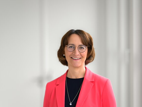 Eine lächelnde Frau mit Brille trägt einen pinkfarbenen Blazer und steht vor einem hellen, unscharfen Hintergrund.