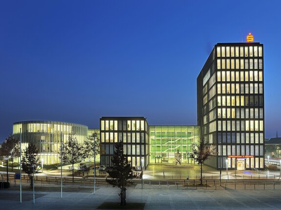 Moderne Gebäude bei Nacht, beleuchtet mit großen Fensterfronten und umgeben von Bäumen und Wegen.