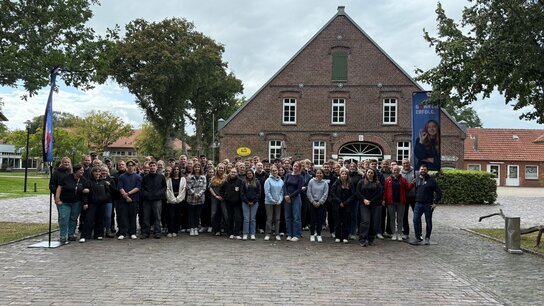 Gruppennfoto von einer großen Menschenmenge vor einem historischen Gebäude in einer ländlichen Umgebung bei bewölktem Himmel.