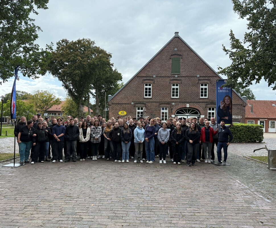 Gruppennfoto von einer großen Menschenmenge vor einem historischen Gebäude in einer ländlichen Umgebung bei bewölktem Himmel.
