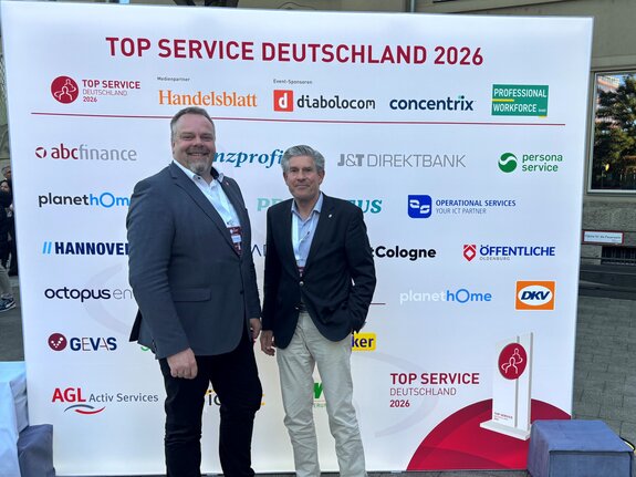 Joachim Köster und Christian Bunk stehen vor einer Werbebande bei Preisverleihung TOP SERVICE 2026