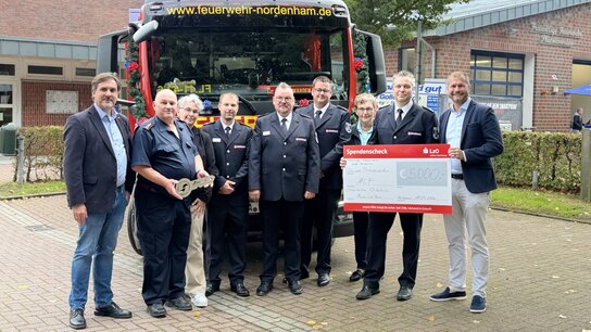 Gruppierung von Feuerwehrmitgliedern und Unterstützern vor einem Feuerwehrfahrzeug, das einen Scheck über 3.000 Euro präsentiert.