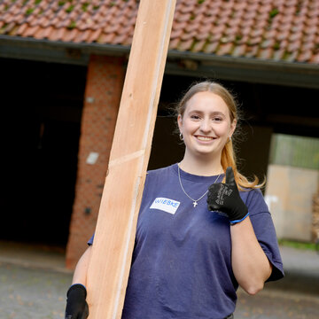 Junge Frau mit Holzlatte, die lächelt und Daumen hoch zeigt, im Freien vor einem Gebäude.