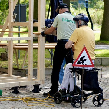Zwei Personen arbeiten an einem Holzgerüst; eine sitzt im Rollator, ein Warnschild weist auf Arbeiten hin.