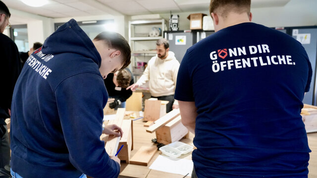 Mehrere Personen arbeiten an einem Tisch mit Holzmaterialien und Werkzeugen. Einige tragen blaue T-Shirts mit Schriftzügen.