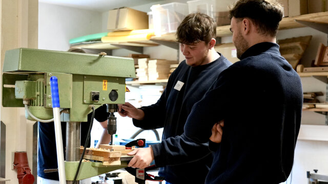 Zwei Personen stehen an einer Bohrmaschine in einer Werkstatt und arbeiten zusammen an einem Holzprojekt.