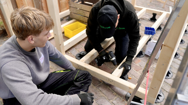 Zwei Personen arbeiten gemeinsam an einem Holzprojekt, während einer ein Teil fixiert und der andere zuschaut.