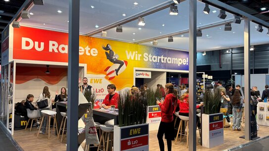 Messestand mit mehreren Personen, Stehtischen und Bannern, die "Du Rakete" und "Wir Startrampe!" zeigen.