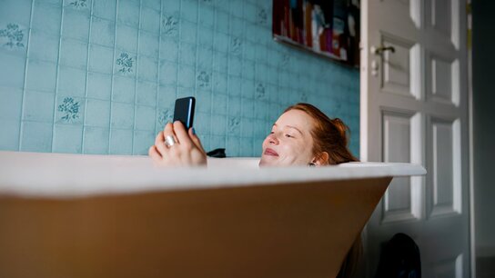 Eine Person in einer Badewanne entspannt sich und hält ein Smartphone in der Hand, umgeben von blauer Fliesenwand.