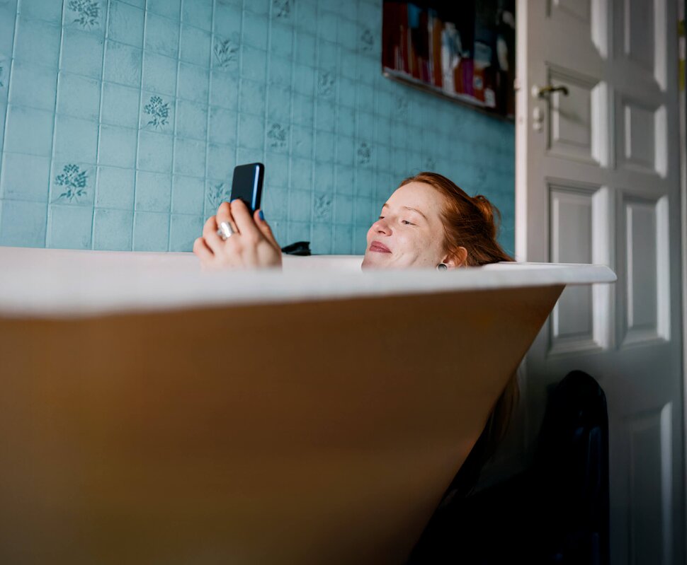 Eine Person in einer Badewanne entspannt sich und hält ein Smartphone in der Hand, umgeben von blauer Fliesenwand.