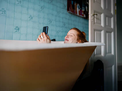 Eine Person in einer Badewanne entspannt sich und hält ein Smartphone in der Hand, umgeben von blauer Fliesenwand.