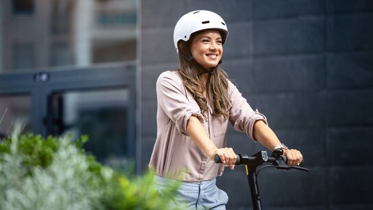 Lächelnde Frau mit Helm fährt auf einem E-Scooter vor einer modernen Gebäude-Fassade. Grüne Pflanzen im Vordergrund.