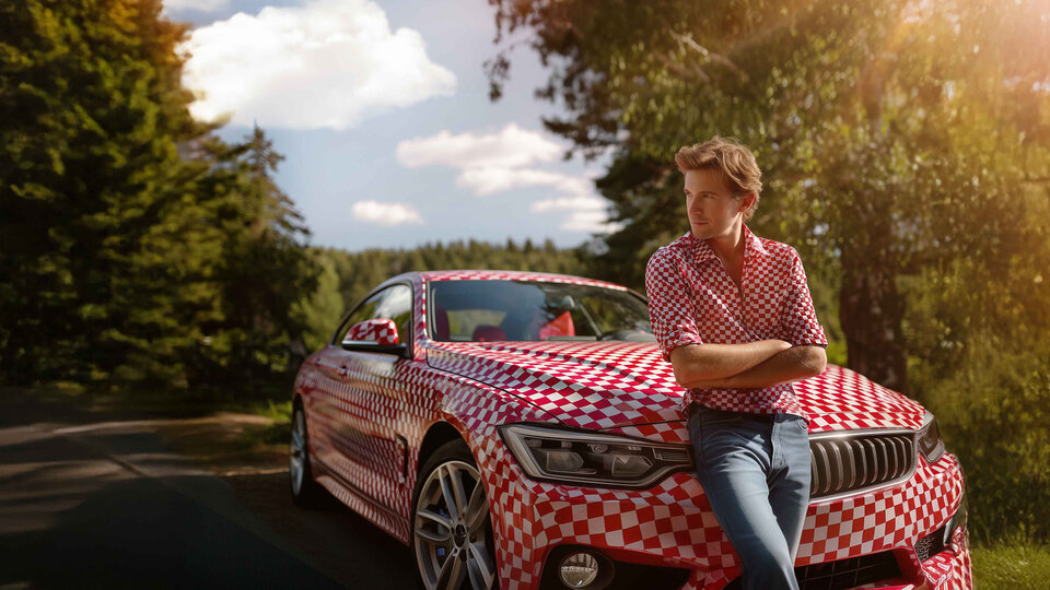 KI-generiertes Bild: Mann in kariertem Hemd lehnt an einem rot-weißen Auto auf einer Waldstraße unter blauem Himmel.