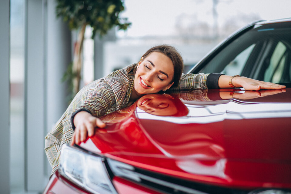 Eine brünette junge Frau in einem karierten Bläser umarmt in einem Auto die Motorhaube von einem roten Auto.
